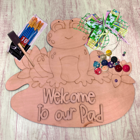 wooden blank kit of frog on lily pad includes 3d letters that say welcome to our pad. also included ribbon to assemble for the bow, wire and beads for the hanger and a pack of paintbrushes.